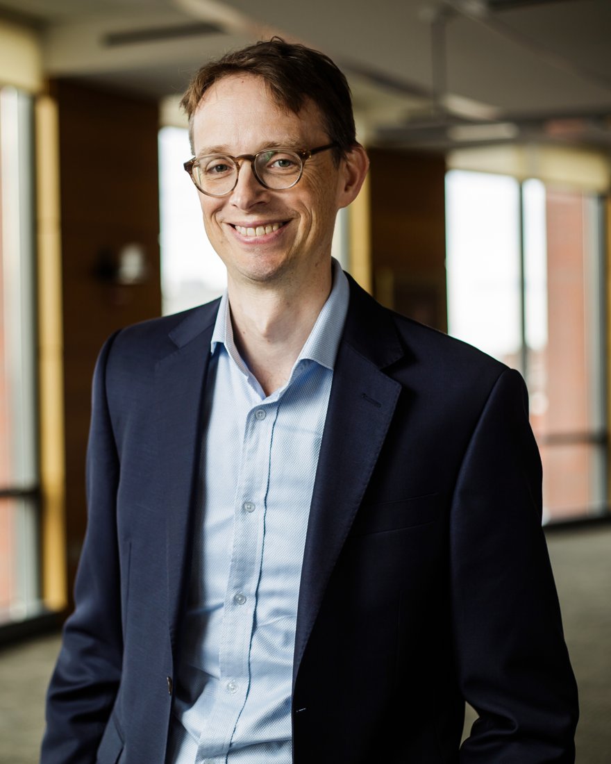 Matthew Bidwell, smiling, in a navy jacket and light blue shirt, standing in a sunlit office.